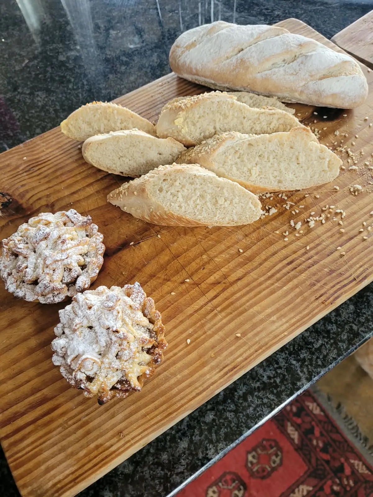 A sliced sesame-topped loaf beside two apple lattice tartlets on a wooden board.