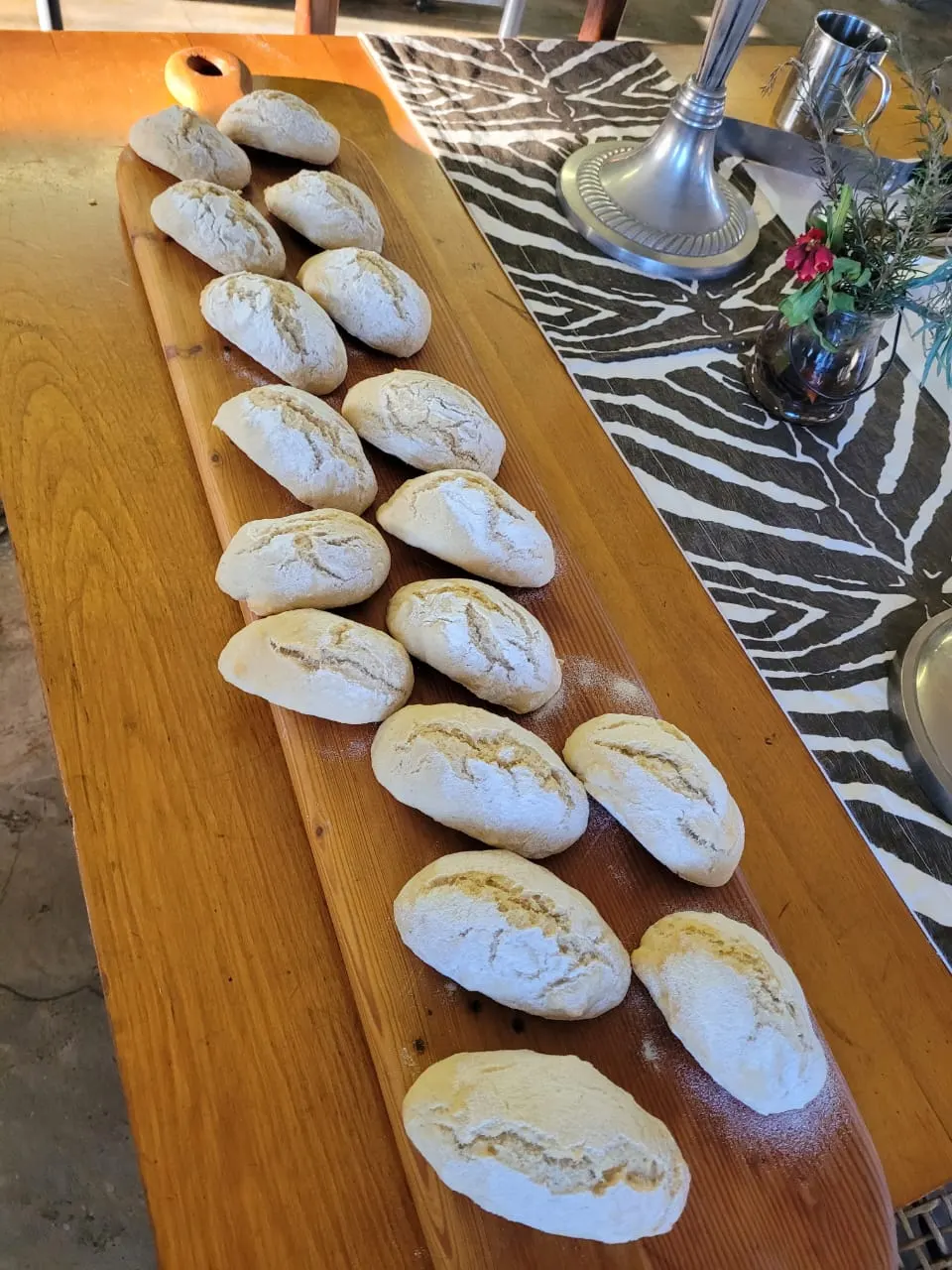 Unbaked, floured bread rolls lined up on a long wooden board.