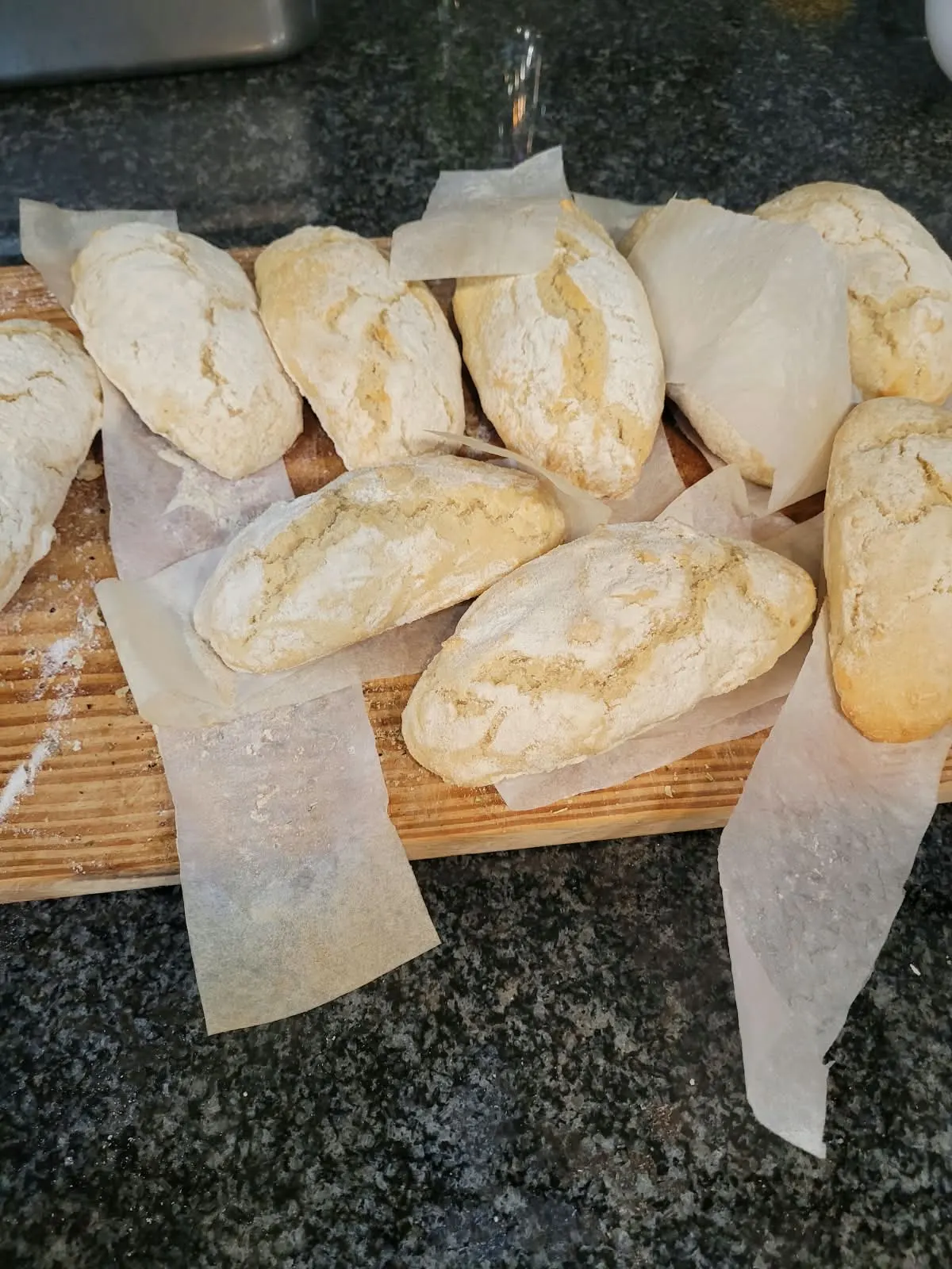 Ten baked bread rolls dusted with flour, laid out on a wooden board.