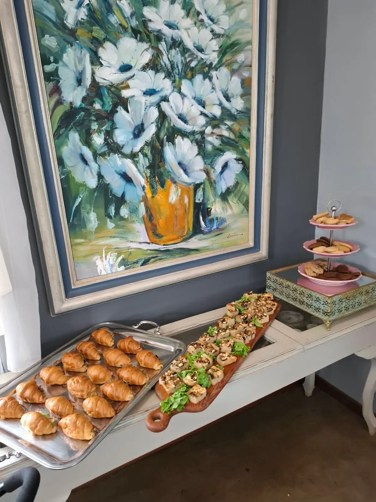 Filled mini croissants, savoury bites and a tier of biscuits beneath a floral painting